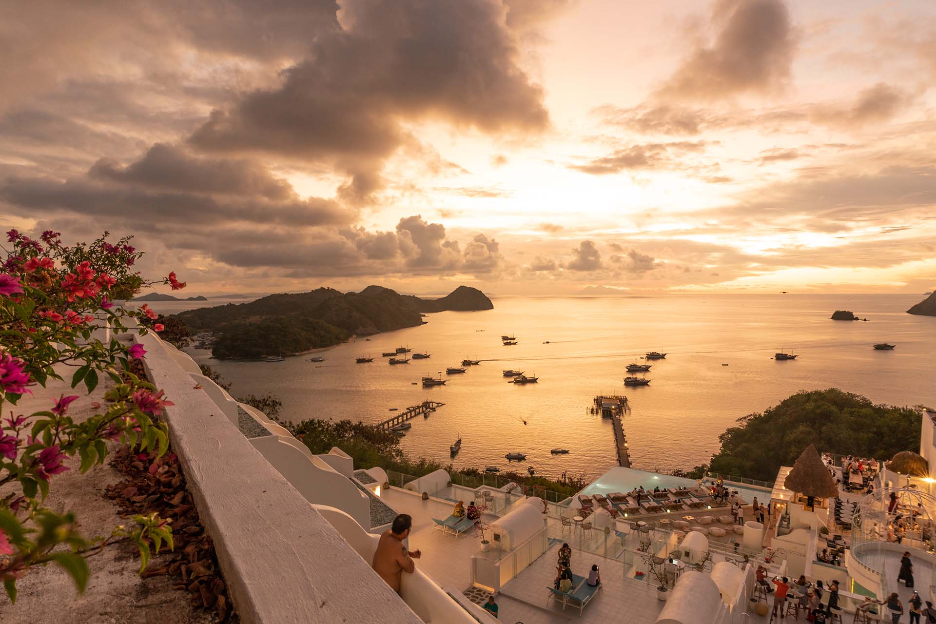 White terraces above Labuan Bajo harbor at sunset, bougainvillea in the foreground and boats resting on the golden water